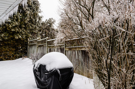 suburban backyard in winter with a snow covered barbecue and ice covered branches, icicles from freezing rain with an overcast sky.の写真素材