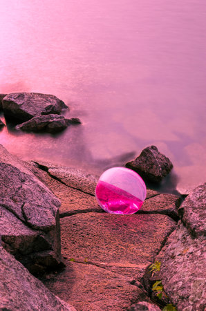 Beach ball trapped on rocks  by waves at the edge of the water under a pink glowing skyの写真素材