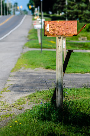 Rusty rural mailbox in front of a row of out of focus mail boxes next to a road in the summer.の写真素材