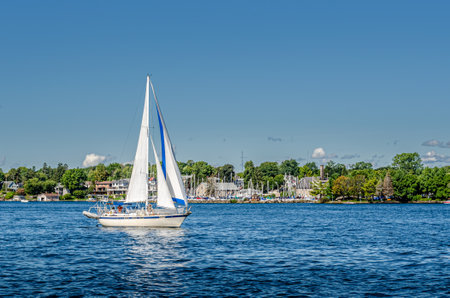 Sailboat sailing past yacht club shoreline with a lightly cloudy sky and blue waterの写真素材