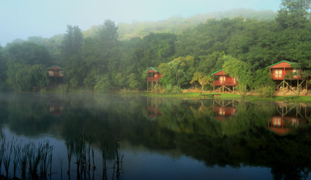 Cosy Log cabins overlooking a lake at Klein-Kariba, in South Africa. The sun is breaking through the fog, illuminating the idyllic setting to have a relaxing surreal holiday.のeditorial素材