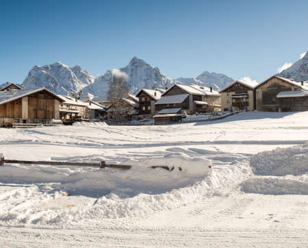 The bright morning sun enlightens a snowy mountain village with farm houses in front of the mountain range of the Piz Lischana groupの写真素材