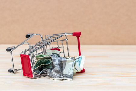 Fallen shopping cart on wooden surface with crumpled up dollar bill. Symbol of consumerism era endの写真素材