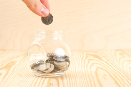 The concept of saving money. Fingers hold a coin directing it to a glass jar with coins on a light...