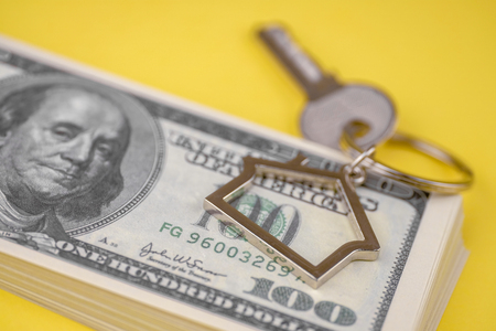 The concept of buying a private house or apartment. Mortgage registration. The accumulation of money for real estate. Metal key chain in the shape of a house with a metal key lying on the pack of hundred US dollars on a yellow background. Close up.の写真素材