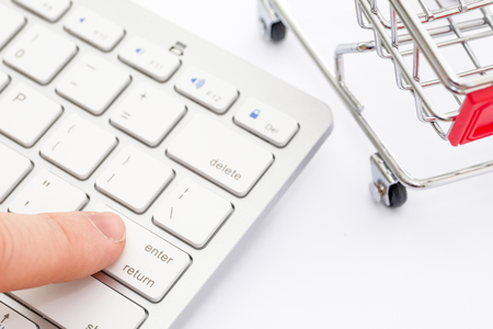 Finger presses the button of the white keyboard on a white background with a cart. The concept of online shopping.の写真素材