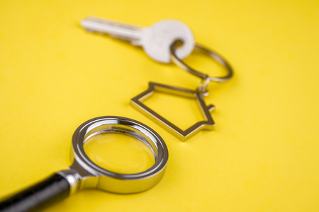 The concept of checking the legal purity of a private house or apartment. Metal keyring in the shape of a house with a metal key under a magnifying glass on a yellow background. Close up.の写真素材