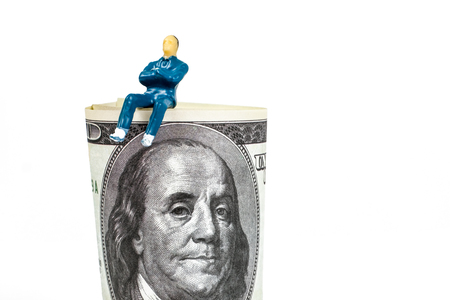 The concept of a successful businessman or stockbroker. A toy man is sitting on a hundred dollar bill against a white background.の写真素材