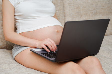 A pregnant girl in a t-shirt works behind a laptop. The concept of employment in the decree.の写真素材
