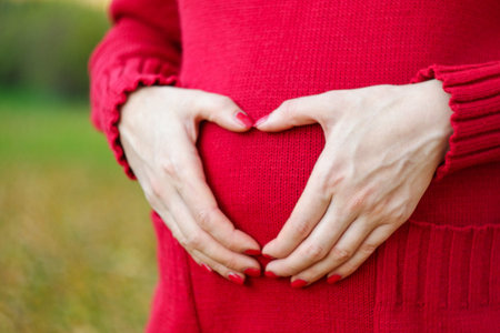 A pregnant girl dressed in a red sweater on the street makes the symbol of love by folding hands...