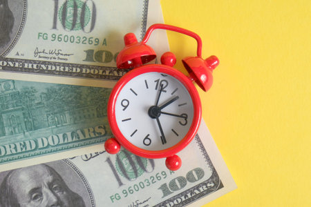 The concept of the start of business investment. A red vintage clock stands on one hundred US dollar bills on a yellow background.の写真素材