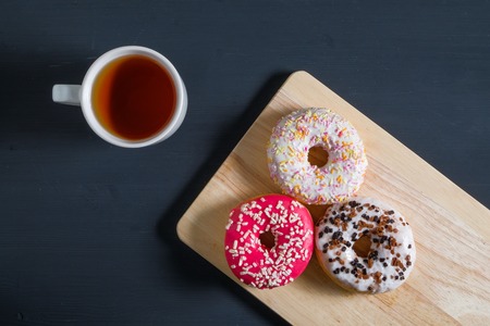 White, pink and brown glazed donuts with cup of tea on black wooden background. Top view.の写真素材