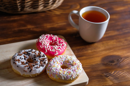 White, pink and brown glazed donuts and cup of tea on wooden background and near rattan basket.の写真素材