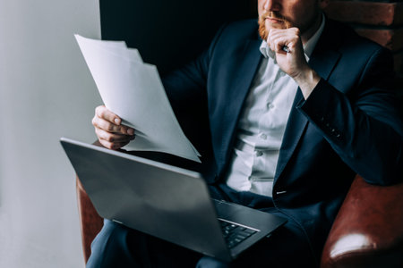 A businessman in a suit sits in an armchair with a laptop and analyzes information from a paper....