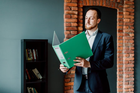 An adult and successful accountant in a suit stands and holds a green folder with reports and considers it in his hands.の写真素材