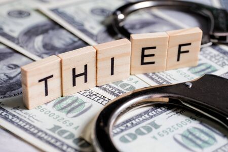 Word made of wooden letters - thief, on a gray background with us dollars next to a magnifier. Crime designation concept. Close up.の写真素材