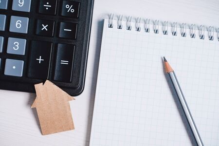 Cardboard cutout house, calculator and pencil on blank sheet of spiral notebook on white background. Concept of construction, budgeting, repairing, property insurance.の写真素材
