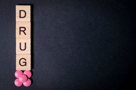 The word drug is on wooden squares next to pink tablets on a black background. Copy space. Top view. Close up.の写真素材