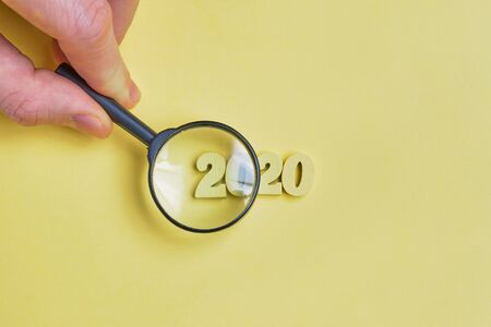 The hand holds a magnifying glass over the abstract year 2020 of wooden digits on a yellow background. Close up.の素材 [FY310137491783]