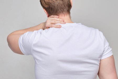 A man stands with his back in a white T-shirt and holds on to his neck because of severe pain....