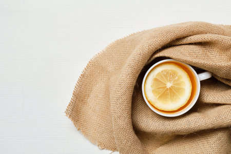 A white cup of tea with lemon in linen on a white wooden background. Copy space. Top view. Close up.