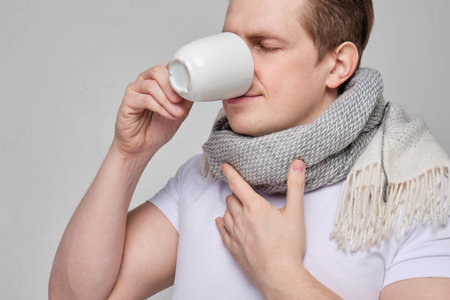 A man in a white T-shirt drinks warm cup of tea from a cold, flu and sore throat. Close up.