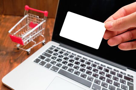 A man holds a card for copy space and mock up against the background of a laptop and a trolley. Close up.の写真素材