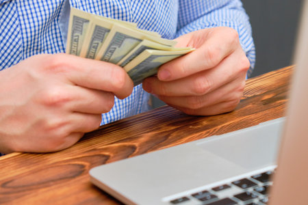 A man counts profit in the form of money from the business in front of the laptop. Close up.