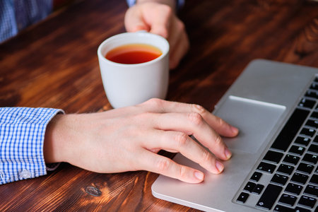 A man drinks tea while working behind a laptop. The concept of rest while working. Close up.