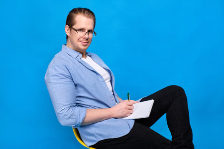 A man in a blue shirt with glasses with a notebook and pen looks at the camera. The concept of a young teacher. Close up.の写真素材