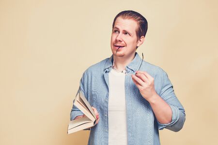 A man in a blue shirt on a yellow background with glasses and holds a book with a thoughtful look. Toned. Close up.の写真素材