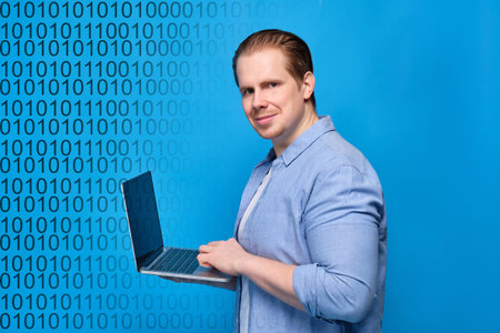 A man in casual clothes on a blue background looks at the camera and programs on a laptop. Binary...