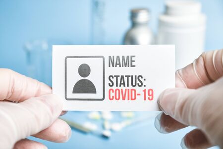 A doctor in gloves holds a white card with the personal data of a person and his status of an infected COVID-19 person against a background of medical equipment and a blue background. Close up.の写真素材