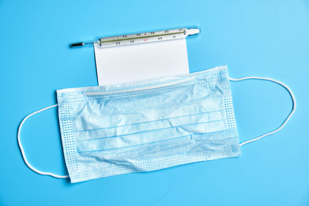 Protective medical face mask. Thermometer and white card on a blue background. Top view.