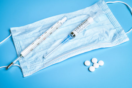 Protective medical face mask and on it is a syringe, thermometer, pills on a blue background. close...