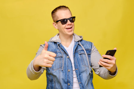 Stylish Caucasian man in a jeans and sunglasses on a yellow background. Holding a smartphone and...
