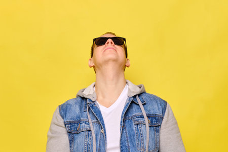 Stylish Caucasian man in a jeans and sunglasses on a yellow background. Copy space. Close up. Raised...