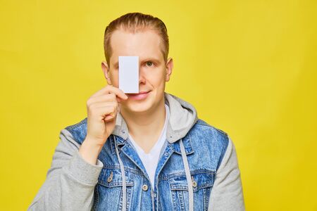 Stylish Caucasian man in a jeans on a yellow background holds a white card at eye level and smiles copy space. Close up.の写真素材
