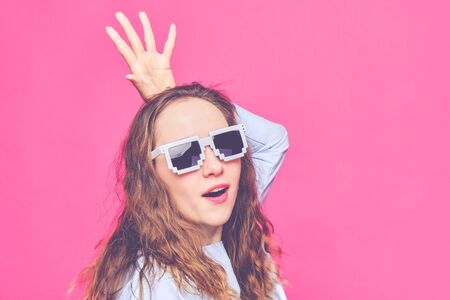 Stylish caucasian girl in a pale blue t-shirt and 8-bit glasses on a pink background. Screaming at the camera and portray the queen. Close up. Toned.の写真素材