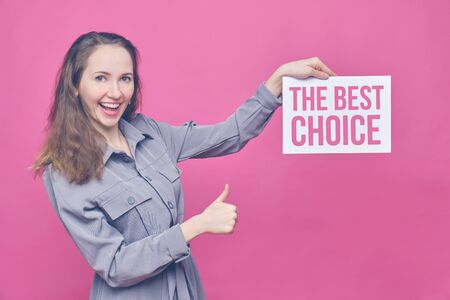 Stylish caucasian girl in a gray dress on a pink background. Hand holds a sheet with white paper and the inscription - the best choice. Close up. Toned.の写真素材