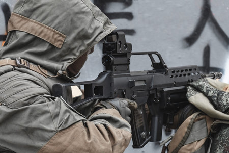 A hired soldier in the hood is aiming from a modern rifle. Close up.