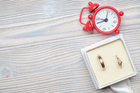 Wedding rings in a box next to the red clock on wooden background. Copy space. Close up.