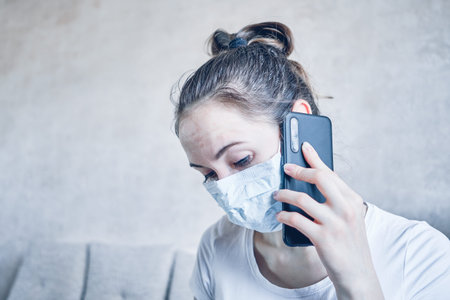 The girl in the medical mask is in quarantine at home and is sadly talking on the smartphone. Toned....