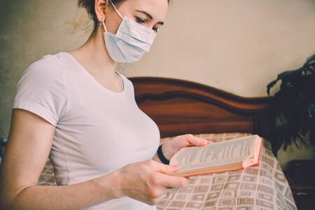 A girl in a medical mask is in quarantine at home and is reading a book. Close up. Toned.の写真素材
