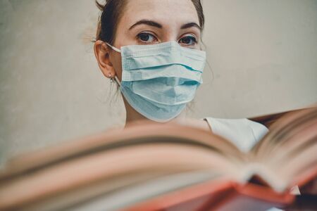 The girl in a medical mask is on self-isolation at home holding a book and looking at the camera. Close up.の写真素材
