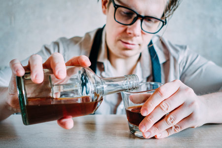 A man in a glasses shirt pours whiskey or cognac into a glass after a hard working day. Close up.の写真素材