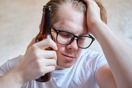 A man in glasses with a bottle of beer rests on his head. Close up.
