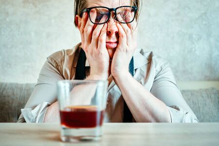 A man in glasses with a glass of whiskey rubs his eyes. Close up.の写真素材