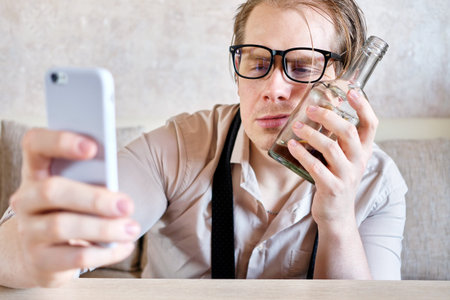 A drunk man in glasses put his face to a bottle of alcohol and looks at the smartphone. Close up.