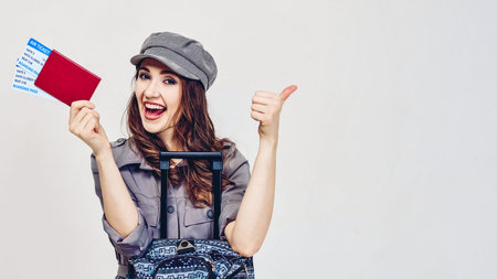 Glamorous young girl in a gray dress holds plane tickets with a passport and shows thumb up. Portrait on a gray background.の写真素材